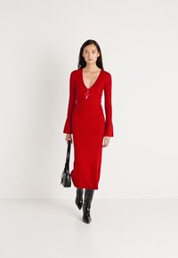 Rød ribbestrikket kjole med v-hals, lange vide ermer og en tettsittende silhuett. Brukt med svarte knelang støvler og en liten svart veske.