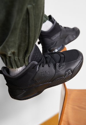 Person trägt schwarze Sportschuhe mit "23" an der Seite, weiße Socken und olivgrüne Hosen und ruht die Füße auf einem Holzstuhl.