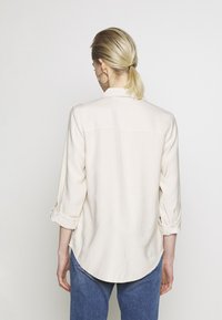 Ljusbeige knapp-up-skjorta med uppvikt ärmar, en spetsig krage och en rundad nederkant. Slät tygtextur, minimala detaljer.