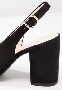 Sandale à talon bloc en daim noir avec bride à boucle argentée et dos ouvert, montrant une partie de la semelle intérieure beige rembourrée.