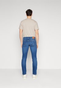 Camiseta ligera de color beige claro para hombre, jeans ajustados azules con bolsillos traseros, parche de cuero en la cintura y zapatillas blancas. Fondo liso.