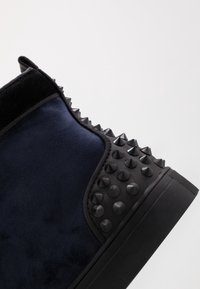 Granatowe zamszowe sneakersy w stylu wysokim z czarnymi ćwiekowanymi akcentami i teksturowaną gumową podeszwą. Zawierają wyściełany kołnierz i gładkie wykończenie.