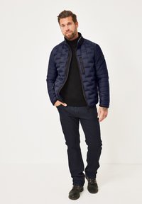 Mann in einer marineblauen Steppjacke, schwarzem Rollkragenpullover, dunklen Jeans und schwarzen Stiefeln, der vor einem einfarbigen hellen Hintergrund steht.