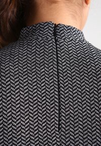 Gros plan sur un haut en tissu texturé noir et blanc avec une fermeture éclair au dos, porté par une personne aux cheveux bruns attachés.