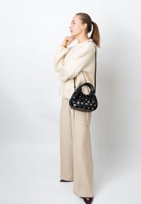 Crna kožna torba sa sferičnim zakovicama, kratkim ručkama i podesivim remenom; model nosi krem džemper i široke bež hlače.