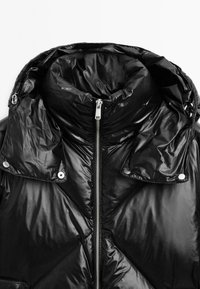 Giacca a vento nera con colletto alto, texture lucida e trapuntata, zip frontale e bottoni a pressione sul colletto. Dettagli di design minimalista.