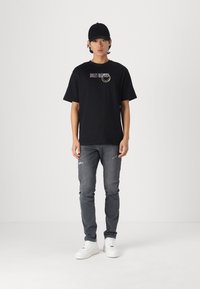 Camiseta de algodón negra con gráfico de "SMILEY", combinada con jeans gris desgastados y zapatillas blancas. El modelo lleva una gorra negra.
