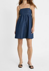 Denimmekko, jossa on ohuet olkaimet, istuva yläosa ja levenevä hame. Tummansininen väri, sileä pinta ja hame ulottuu polven yläpuolelle.