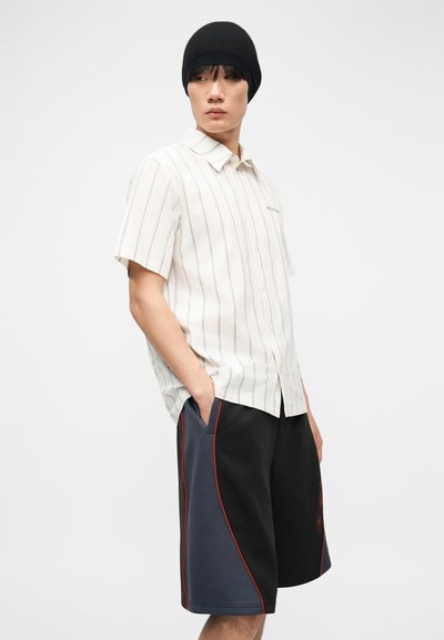 Νέος άνδρας που φοράει μαύρο σκουφί, άσπρο ριγέ πουκάμισο με κοντά μανίκια και μαύρο-γκρι σορτς με κόκκινη διακόσμηση, στέκεται με τα χέρια στις τσέπες.