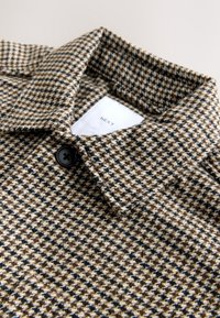Manteau à col avec motif pied-de-poule en marron, noir et beige, avec un bouton noir unique et une étiquette blanche avec le texte "NEXT".