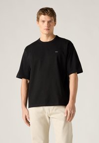 Μαύρο κοντομάνικο t-shirt από βαμβακερό ύφασμα, με στρογγυλή λαιμόκοψη και μικρό ανάγλυφο λογότυπο στο στήθος. Φο ρεμένο με ανοιχτόχρωμο τζιν.