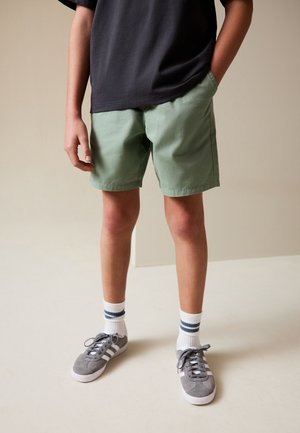 Barn iført grønne shorts, sort t-shirt, hvide sokker med blå striber og grå sneakers, stående med den ene hånd i lommen mod en ensfarvet baggrund.