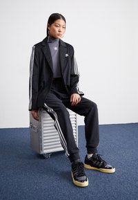 Blazer preto com riscas brancas nas mangas, suéter cinza de gola alta, calças de moletom pretas com riscas e ténis pretos com solas creme.
