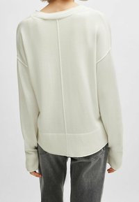 Ljust krämfärgad tröja med oversized passform, ribbad halsringning och ärmslut, sömlösa axlar och rundad nederkant, tillsammans med mörkgrå jeans.