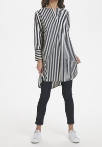 Túnica a rayas en azul marino y crema con cuello alto, cierre frontal con botones, mangas largas y dobladillo curvo, combinada con vaqueros oscuros y zapatillas blancas.