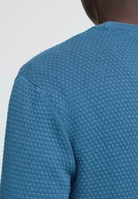 Struktura u plavoj boji pletenog pulovera s okruglim izrezom, s uzorkom nalik na vafle i rebrastim detaljima na izrezu i ramenima.