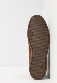 Semelle d'une chaussure en suède marron avec un motif strié diagonal et des logos, positionnée verticalement contre un coin blanc en arrière-plan.