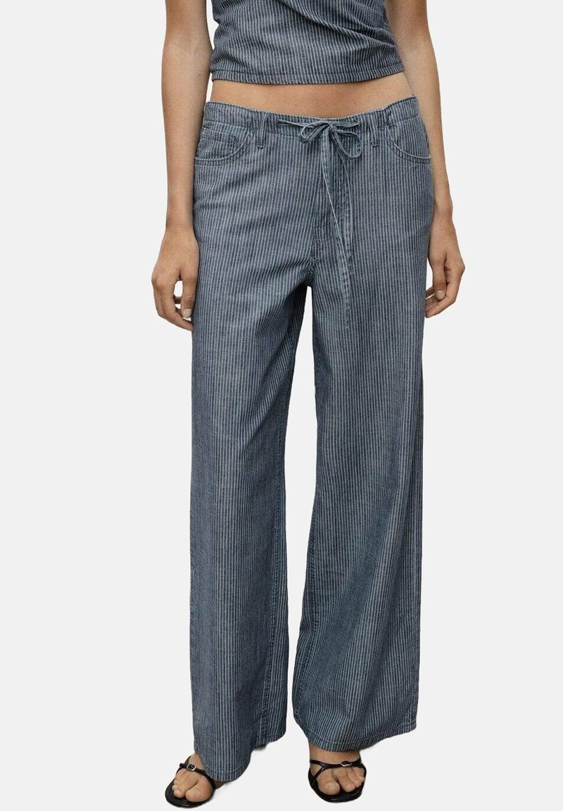 Personne portant un pantalon à rayures verticales bleu et blanc, taille haute, coupe ample avec taille à cordon et sandales noires ouvertes.