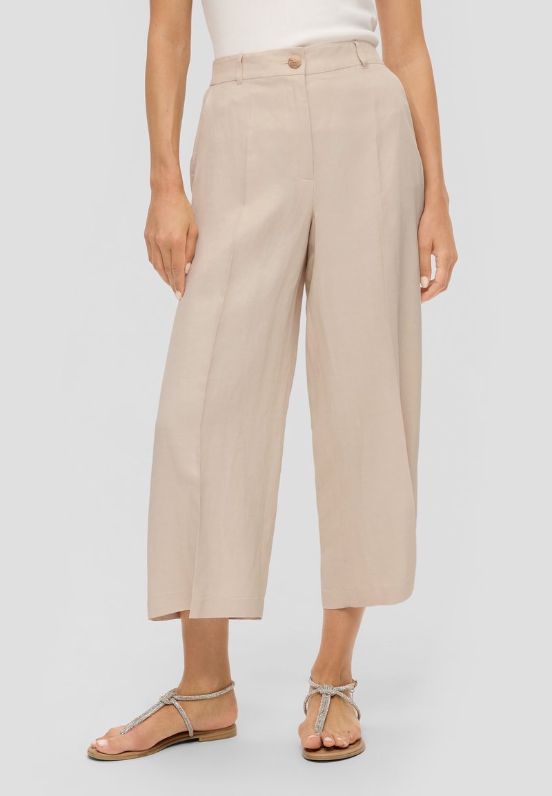 Pantalon large beige taille haute en tissu léger, avec un bouton à l'avant et deux poches latérales. Associé à des sandales argentées.