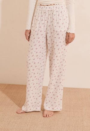 Personne debout pieds nus portant un pantalon blanc ample avec un petit motif floral rose et un haut blanc à manches longues avec des bords festonnés.