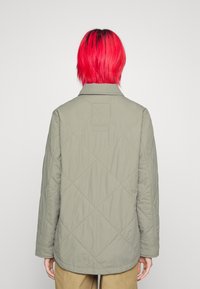 Vadderad olivgrön jacka med krage, långa ärmar och en bakre ok. Har diamantstickning och en fyrkantig lapp på baksidan.