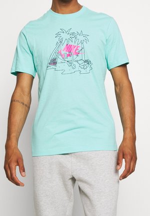 Turkoosi puuvillapaita, jossa on graafinen suunnittelu vaaleanpunaisilla yksityiskohdilla, palmuja ja tyylitelty Nike-logo. Lyhyet hihat, pyöreä kaula-aukko.