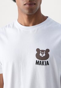 Bílá bavlněná tričko s grafikou hnědého medvěda a nápisem "MAKIA" v oblasti levého prsu. Kulatý výstřih, krátké rukávy.