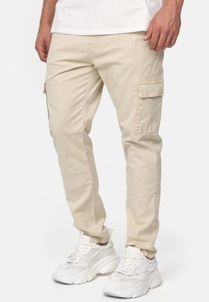 Pantalon cargo beige avec poches à rabat latérales porté avec des baskets blanches épaisses et une chemise blanche, montré de la mi-cuisse jusqu'aux pieds.