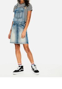 Denim overall-mekko vaaleansinisellä pesulla, etutasku, säädettävät olkaimet, yhdistetty raidalliseen paitaan ja mustiin korkeisiin tennareihin.