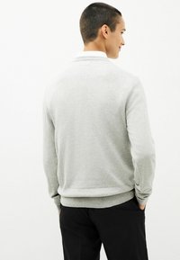 Lys grå sweater med en rund krave, ribbede manchetter og kant. Glat tekstur, tætsiddende design, båret over sorte bukser, set fra ryggen.