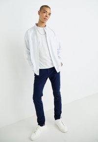 Une chemise à rayures blanches et bleu clair, boutonnière, portée sur un t-shirt blanc, associée à un pantalon bleu foncé et des baskets blanches. Design simple et coupe décontractée.
