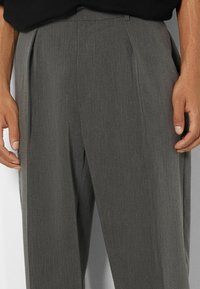 Man som bär grå skräddarsydda byxor med veck, står med händerna avslappnat vid sidorna mot en neutral bakgrund.