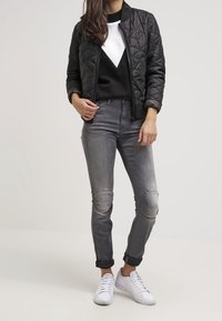 Prošívaná černá bomber bunda přes černobílou mikinu, doplněná šedými slim-fit džíny a bílými teniskami.