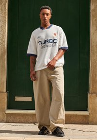 White oversized short-sleeve jersey with blue accents, graphic "TURBO" text, beige wide-leg pants, and black shoes, set against a green door.