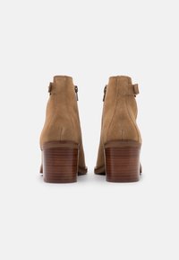 Botas de tobillo de ante marrón con un tacón de bloque de madera, cremallera trasera y detalle de correa ajustable en el tobillo. Textura suave y diseño estilizado.