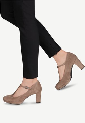 Brązowe zamszowe buty na obcasie z designem T-strap, okrągłym noskiem i ozdobnym przeszyciem wzdłuż krawędzi. Założone z czarnymi zwężającymi się spodniami.