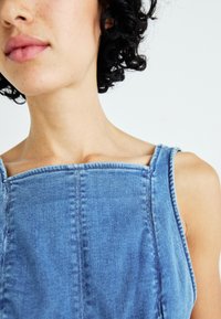 Jeansjurk met een vierkante halslijn, verstelbare schouderbanden en zichtbare stiksels. Lichtblauwe kleur met een gladde textuur.