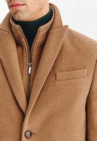 Brązowy płaszcz z wełny z dodatkiem, o fakturowanym wykończeniu, z kołnierzykiem notch, zapięciem na zamek błyskawiczny i kieszenią na piersi. Nałożony na zielony golf.