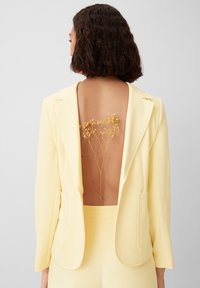 Yellow blazer with a deep back cutout featuring dried yellow flowers. Smooth fabric, tailored design, and dual front pockets.