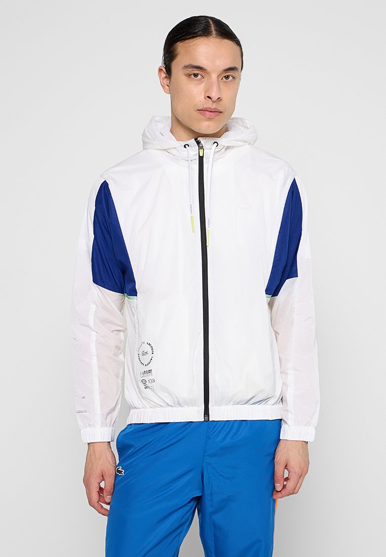 Veste légère blanche avec des manches bleues, capuche ajustable et fermeture éclair à l'avant ; présente un logo imprimé et une taille élastique ; texture lisse.
