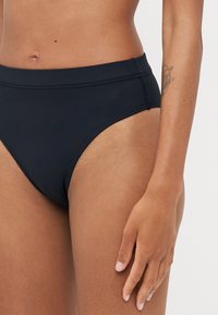 High-waisted bikini bottom in black fabric, smooth texture, with a simple design and clean lines, featuring contrasting stitching.