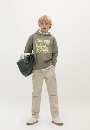 Felpa con cappuccio verde oliva con grafica gialla, abbinata a pantaloni cargo beige e scarpe da ginnastica bianche. Porta una borsa verde scuro.
