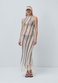 Robe au crochet crème avec des rayures marron, décolleté en halter et ourlet frangé. Associée à une casquette beige et des tongs noires.