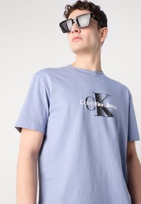 Hele lavendelvangust t-särk lühikeste käiste ja ümmarguse kaelusega, mille esiosal on musta ja valge värvi "Calvin Klein Jeans" logo.