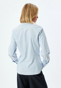 Vaaleansininen nappikauluspaita, jossa on rakenteellinen kaulus, slim fit -muotoilu ja pitkät hihat, yhdistettynä viistopintaisiin housuihin. Sileä pinta.