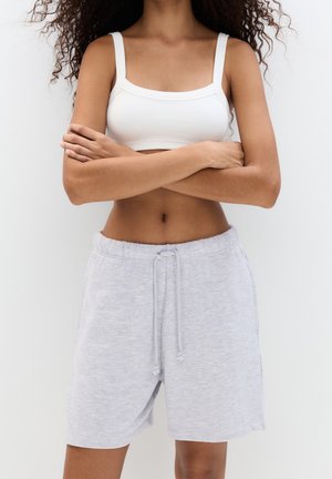 Kobieta z założonymi skrzyżowanymi rękami, ubrana w biały krótki top i jasnoszare szorty ze ściągaczem na tle jednolitego tła.