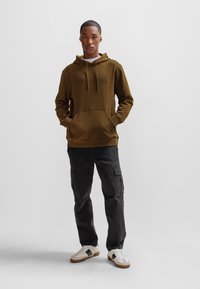 Felpa verde oliva con tasca frontale, cappuccio con cordini e patch con logo. Abbinata a pantaloni cargo scuri e sneakers bianche con dettagli marroni.