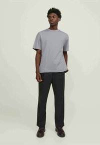 Homem em pé, vestindo uma t-shirt cinza simples de mangas curtas, calças pretas e sapatos castanhos-escuros, em frente a um fundo claro e liso.