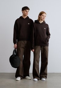 Δύο άτομα φορούν όμοιες σκούρες καφέ κουκούλες και παντελόνια καφέ με φαρδιά πόδια. Ένας κρατά μια μαύρη κυκλική τσάντα και φοράει λευκά παπούτσια.