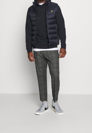 Uomo che indossa un gilet imbottito blu navy, felpa con cappuccio nera, maglietta bianca, pantaloni grigi a quadri e sneaker bianche Nike, in piedi davanti a uno sfondo semplice.